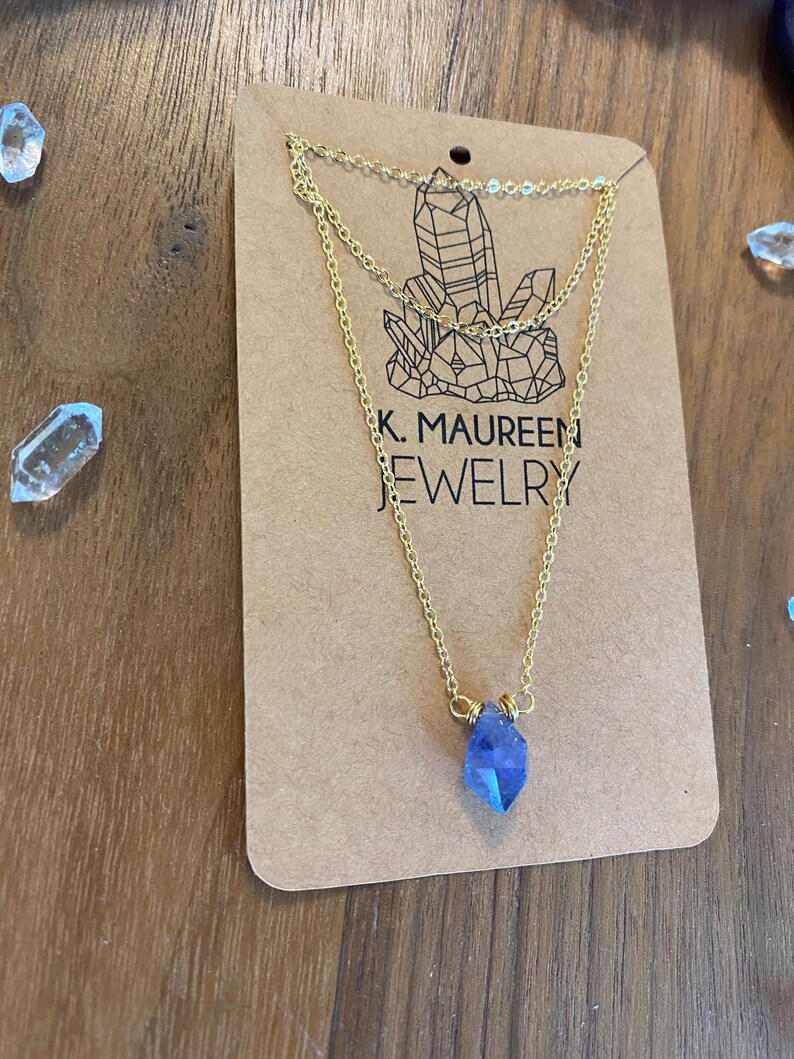 May include: A gold chain necklace with a blue crystal pendant. The necklace is layered and has a delicate design. The pendant is a faceted crystal with a pointed shape. The necklace is displayed on a brown card with the text "K. Maureen Jewelry".