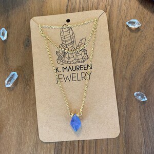 May include: A gold layered chain necklace with a purple amethyst crystal pendant. The necklace is displayed on a brown card with the text "K. Maureen Jewelry".