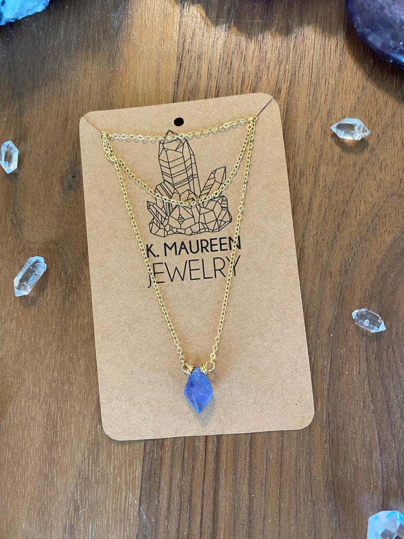 May include: A gold layered chain necklace with a purple amethyst crystal pendant. The necklace is displayed on a brown card with the text "K. Maureen Jewelry".