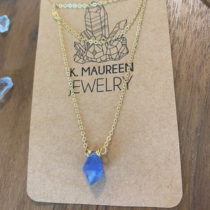 May include: A delicate gold chain necklace with a light blue crystal pendant. The necklace is displayed on a brown card with the text "K. Maureen Jewelry".