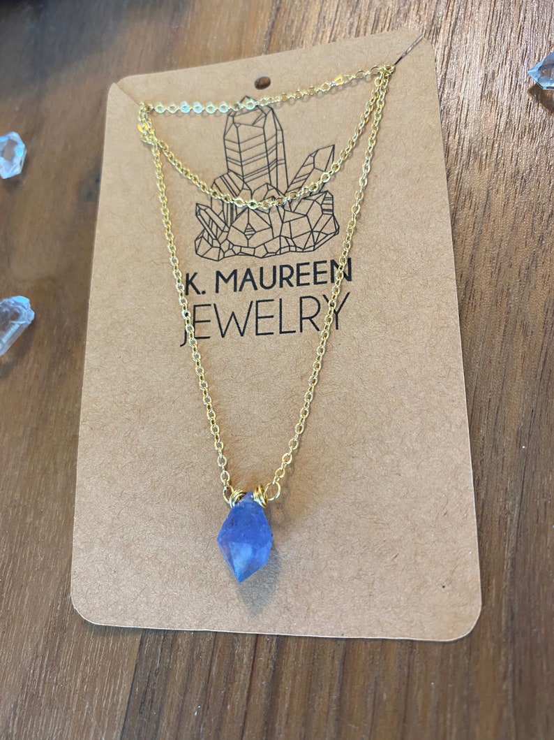 May include: A delicate gold chain necklace with a light blue crystal pendant. The necklace is displayed on a brown card with the text "K. Maureen Jewelry".