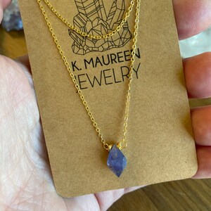 May include: A gold chain necklace with a small, faceted, purple amethyst crystal pendant. The necklace is on a brown card with the text "K. MAUREEN JEWELRY".