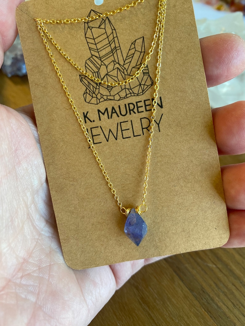 May include: A gold chain necklace with a small, faceted, purple amethyst crystal pendant. The necklace is on a brown card with the text "K. MAUREEN JEWELRY".