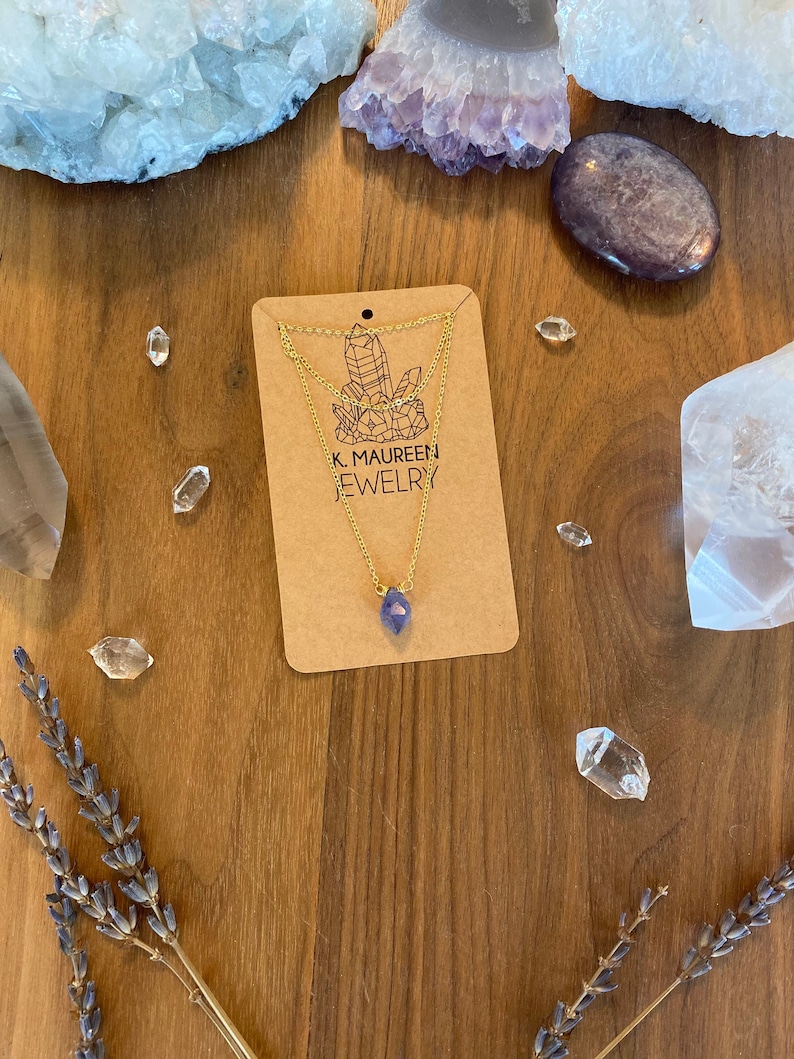 May include: A gold chain necklace with a purple gemstone pendant. The necklace is displayed on a brown wooden surface with crystals and lavender sprigs. The card reads "K. Maureen Jewelry".