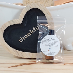 May include: A small clear plastic bag containing a brown glass bottle with a black cap. The bottle is labeled "SLEEP TIGHT" and contains a blend of essential oils. The bag is sitting on a wooden surface with a heart-shaped sign that says "thankful".