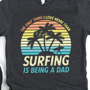 Pu&ograve; includere: Una maglietta grigia scura con una grafica di tramonto retr&ograve; e il testo "L'unica cosa che amo pi&ugrave; del surf &egrave; essere pap&agrave;."
