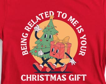 May include: A red t-shirt with a festive Christmas design. The graphic shows a cartoon Christmas tree and a gift box with arms and legs, set against a yellow circle. The text reads, "BEING RELATED TO ME IS YOUR CHRISTMAS GIFT."