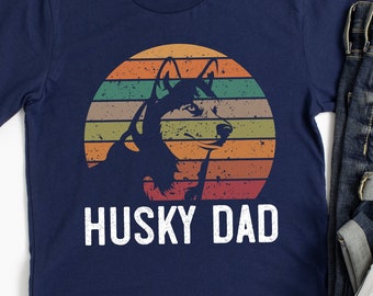Peut inclure: Un t-shirt bleu marine avec un graphique de style vintage d'un chien husky devant un coucher de soleil. Le texte "Husky Dad" est imprimé en blanc sous le graphique.