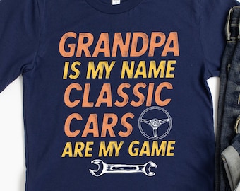 Puede incluir: Una camiseta azul marino con el texto "Grandpa is my name Classic Cars are my game" en naranja y amarillo. También se incluye un volante y una llave inglesa en el diseño.