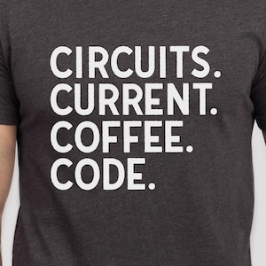 Könnte beinhalten: Dunkelgraues T-Shirt mit dem weißen Aufdruck "CIRCUITS. CURRENT. COFFEE. CODE.". Der Text ist vertikal angeordnet. Das T-Shirt hat einen Rundhalsausschnitt und kurze Ärmel.