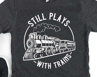 May include: A dark gray t-shirt with a white graphic of a train and the text "Still Plays With Trains".