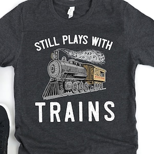 May include: A dark gray t-shirt with the text "Still Plays With Trains" in white. A vintage-style illustration of a steam train with a passenger car is centered on the shirt.