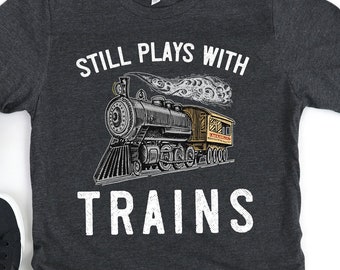 May include: A dark gray t-shirt with the text "Still Plays With Trains" in white. A vintage-style illustration of a steam train with a passenger car is centered on the shirt.