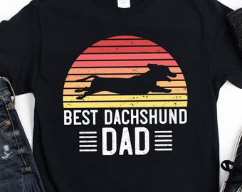 Può includere: Maglietta nera con una grafica di tramonto retrò e una silhouette di un bassotto. Il testo "Best Dachshund Dad" è stampato sotto la grafica.