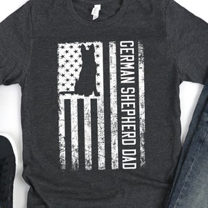 May include: A dark grey t-shirt with a distressed white American flag design and the text "GERMAN SHEPHERD DAD". The flag design includes a silhouette of a German Shepherd dog.