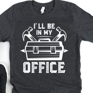 Könnte beinhalten: Ein dunkelgraues T-Shirt mit einer weißen Grafik einer Werkzeugkiste mit zwei Hämmern und dem Text "Ich bin in meinem Büro".