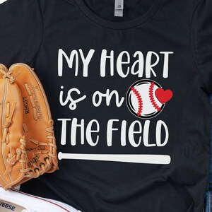 Könnte beinhalten: Schwarzes T-Shirt mit dem Text "My heart is on the field" und einer Baseballgrafik. Das Shirt liegt auf einer weißen Oberfläche mit einem Baseballhandschuh, einem Baseball und einem Paar weißen Sneakers.