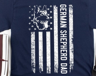 May include: A navy blue t-shirt with a distressed American flag design, a German Shepherd dog, and the text "GERMAN SHEPHERD DAD" in white. The image includes dark grey trousers and maroon high-top trainers.
