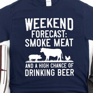 May include: A navy blue t-shirt with white text that reads "Weekend Forecast: Smoke Meat and a High Chance of Drinking Beer". The shirt features a cartoon cow, pig, and chicken.