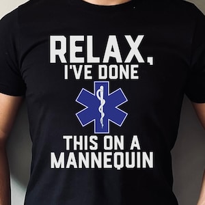 Könnte beinhalten: Schwarzes T-Shirt mit weißem Text: "RELAX, I'VE DONE THIS ON A MANNEQUIN." Ein blaues medizinisches Symbol ist mittig auf dem T-Shirt. Das T-Shirt hat kurze Ärmel.