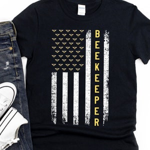 Pode incluir: Camiseta preta com um design de bandeira americana branca e amarela. A bandeira tem uma fileira de abelhas no topo e a palavra "BEEKEEPER" verticalmente para baixo no lado direito.