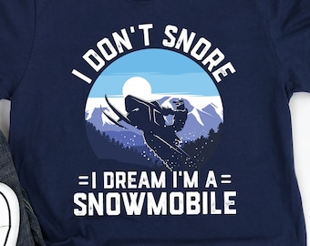 Puede incluir: Una camiseta azul marino con un gráfico blanco de una moto de nieve en un paisaje de montaña nevado. El texto en la camiseta dice "I don't snore = I dream I'm a snowmobile".