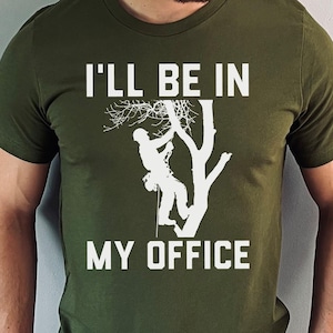 以下が含まれることがあります： オリーブグリーンのTシャツ。白い文字で「I'LL BE IN MY OFFICE」と、木登りをする人のグラフィックが描かれています。木登りをする人は木のシルエットです。Tシャツは半袖です。