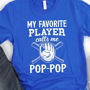 May include: A royal blue t-shirt with white text that reads "My Favorite Player calls me Pop-Pop". The text is accompanied by a graphic of two crossed baseball bats and a baseball glove.