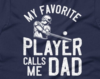 May include: Navy blue t-shirt with a white graphic of a baseball player swinging a bat. The text "My Favorite Player Calls Me Dad" is printed on the shirt.