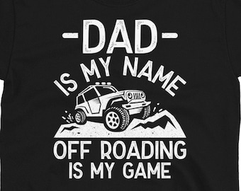Puede incluir: Camiseta negra con texto blanco que dice "Dad is my name, off-roading is my game". Se muestra una silueta blanca de un jeep conduciendo fuera de la carretera.