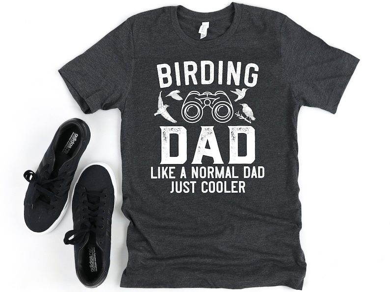 Op de afbeelding: Donkergrijs T-shirt met witte tekst die "Birding Dad Like a Normal Dad Just Cooler" luidt. Het shirt heeft ook een verrekijker en vogels.