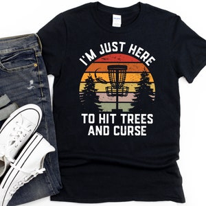 May include: Black t-shirt with a retro sunset graphic and the text "I'm Just Here To Hit Trees And Curse". The graphic features a disc golf basket.