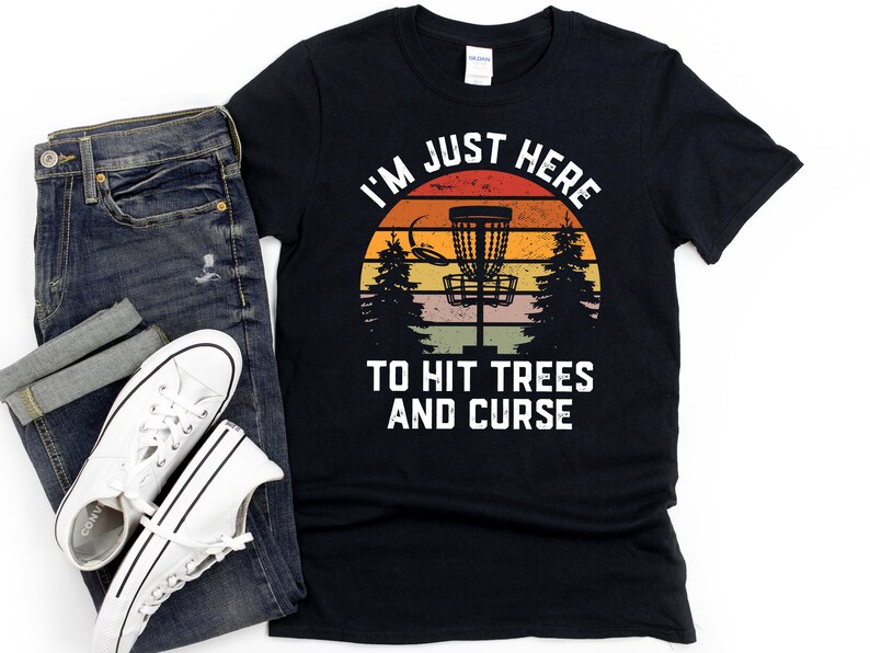 May include: Black t-shirt with a retro sunset graphic and the text "I'm Just Here To Hit Trees And Curse". The graphic features a disc golf basket.