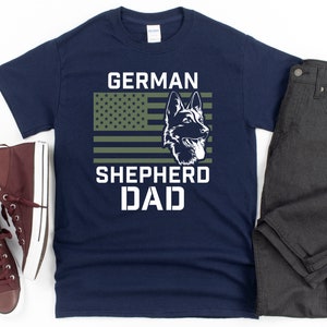 K&ouml;nnte beinhalten: Marineblaues T-Shirt mit einem gr&uuml;nen, wei&szlig;en und roten amerikanischen Flaggen-Design. Die Flagge hat eine Silhouette eines Deutschen Sch&auml;ferhundes in der Mitte. Der Text "GERMAN SHEPHERD DAD" ist unter der Flagge gedruckt.