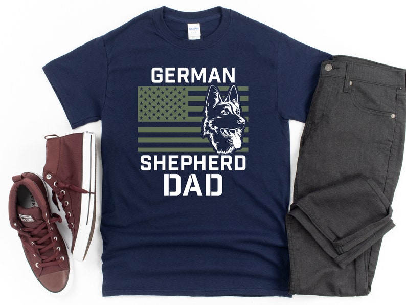 K&ouml;nnte beinhalten: Marineblaues T-Shirt mit einem gr&uuml;nen, wei&szlig;en und roten amerikanischen Flaggen-Design. Die Flagge hat eine Silhouette eines Deutschen Sch&auml;ferhundes in der Mitte. Der Text "GERMAN SHEPHERD DAD" ist unter der Flagge gedruckt.