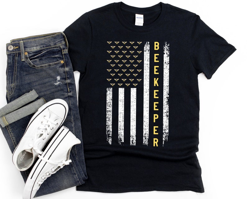 Pode incluir: Camiseta preta com um design de bandeira americana branca e amarela. A bandeira tem uma fileira de abelhas no topo e a palavra "BEEKEEPER" verticalmente para baixo no lado direito.