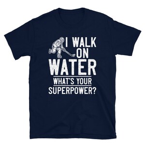 May include: A navy blue t-shirt with a white graphic of a hockey player skating on water. The text "I walk on water" and "What's your superpower?" is printed in white on the shirt.