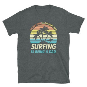 Pu&ograve; includere: Una maglietta grigio scuro melange con una grafica di tramonto con palme e un surfista. Il testo recita "L'unica cosa che amo pi&ugrave; del surf &egrave; essere pap&agrave;."