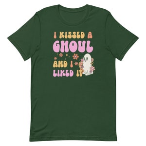 Op de afbeelding: Een groen T-shirt met de tekst "I kissed a ghoul and I liked it" in roze en oranje letters. Naast de tekst staat een cartoon-spook met roze bloemen.