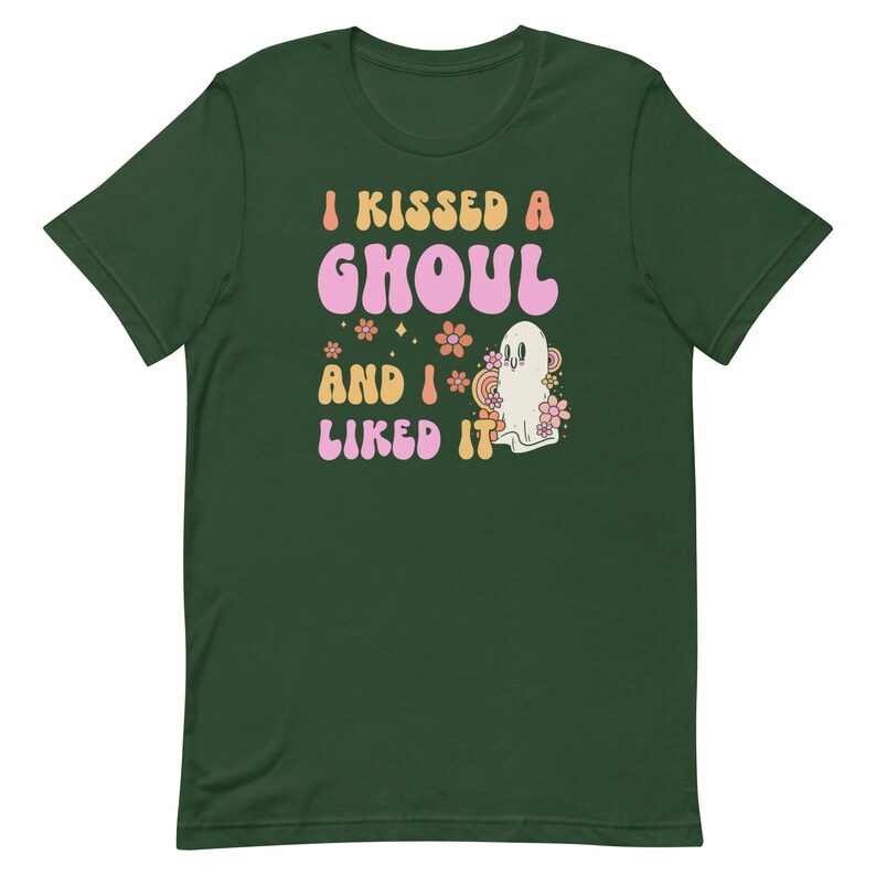 Op de afbeelding: Een groen T-shirt met de tekst "I kissed a ghoul and I liked it" in roze en oranje letters. Naast de tekst staat een cartoon-spook met roze bloemen.