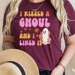 Op de afbeelding: Een maroon t-shirt met de tekst "I kissed a ghoul and I liked it" in een retro lettertype. De tekst is in roze, oranje en geel. Een cartoon-spook met een roze bloem erachter staat ook op het shirt.