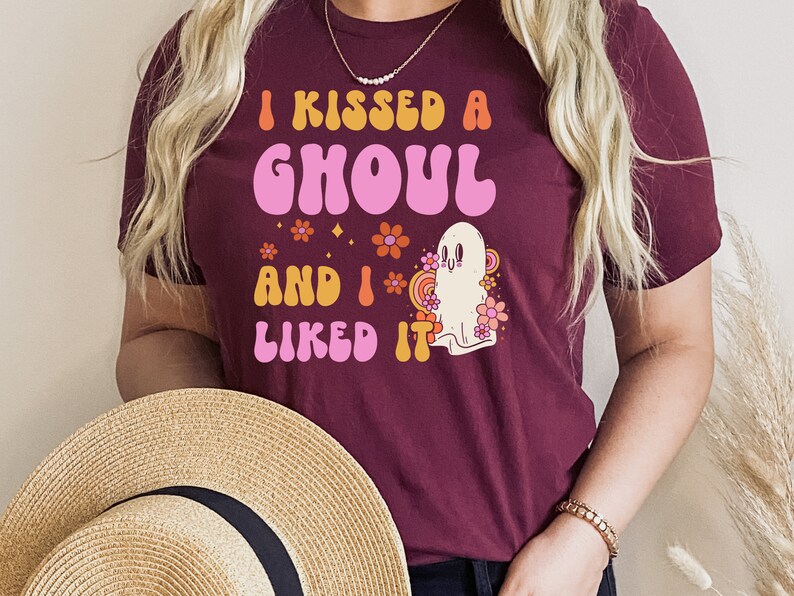 Op de afbeelding: Een maroon t-shirt met de tekst "I kissed a ghoul and I liked it" in een retro lettertype. De tekst is in roze, oranje en geel. Een cartoon-spook met een roze bloem erachter staat ook op het shirt.