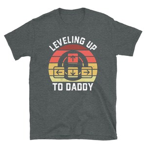 May include: A dark gray t-shirt with a retro-style graphic that says "Leveling Up To Daddy" with an arrow key design.