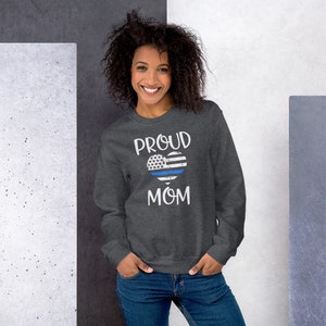 May include: A dark gray sweatshirt with the words "PROUD MOM" in white. The design features a heart-shaped American flag with a blue line. The sweatshirt is made of a soft material.