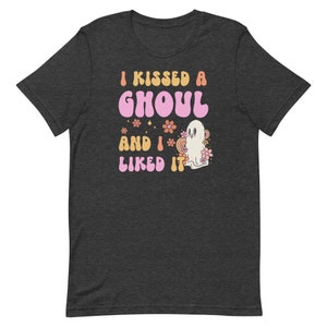 Op de afbeelding: Een donkergrijs T-shirt met een roze en oranje afbeelding waarop staat "I kissed a ghoul and I liked it". De afbeelding bevat ook een geest en bloemen.