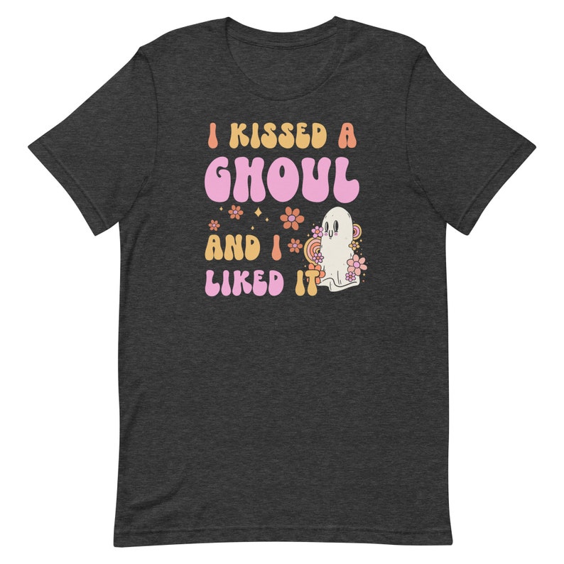 Op de afbeelding: Een donkergrijs T-shirt met een roze en oranje afbeelding waarop staat "I kissed a ghoul and I liked it". De afbeelding bevat ook een geest en bloemen.