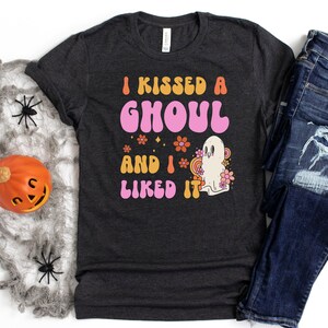 Op de afbeelding: Een donkergrijs T-shirt met de tekst "I kissed a ghoul and I liked it" in roze en oranje letters. Naast de tekst staat een cartoon-spook met bloemen.