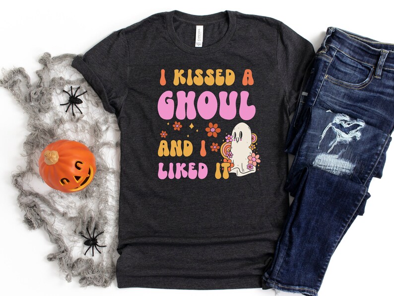 Op de afbeelding: Een donkergrijs T-shirt met de tekst "I kissed a ghoul and I liked it" in roze en oranje letters. Naast de tekst staat een cartoon-spook met bloemen.