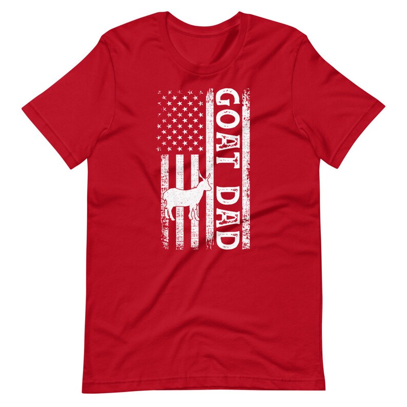 以下が含まれることがあります： 赤いTシャツに、くたびれたアメリカ国旗のデザインと白い文字で「GOAT DAD」と書かれている。国旗の中央には、白いシルエットのヤギが描かれている。