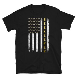 Pode incluir: Camiseta preta com um design de bandeira americana branca e dourada e a palavra "BEEKEEPER" verticalmente do lado direito da bandeira.